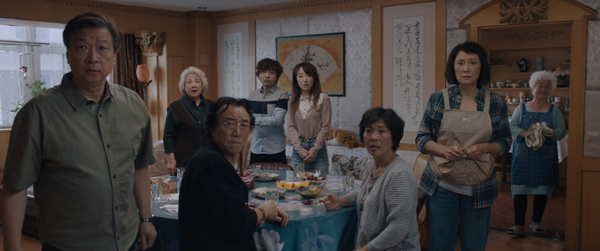 Still from the film "The Farewell" shoes a family of 8 gathered around a large table for a meal. They all look expectantly at the camera, some turning around in their chairs to stare.