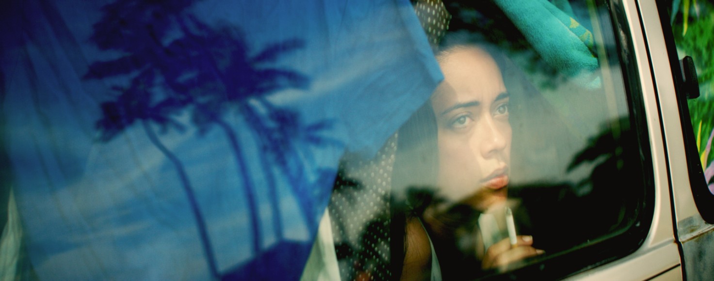 A young person looks out of a car window, seeming to be drinking a fast food drink from a straw, while in the reflection of the window we can see what she may be looking at — palm trees against the blue sky.