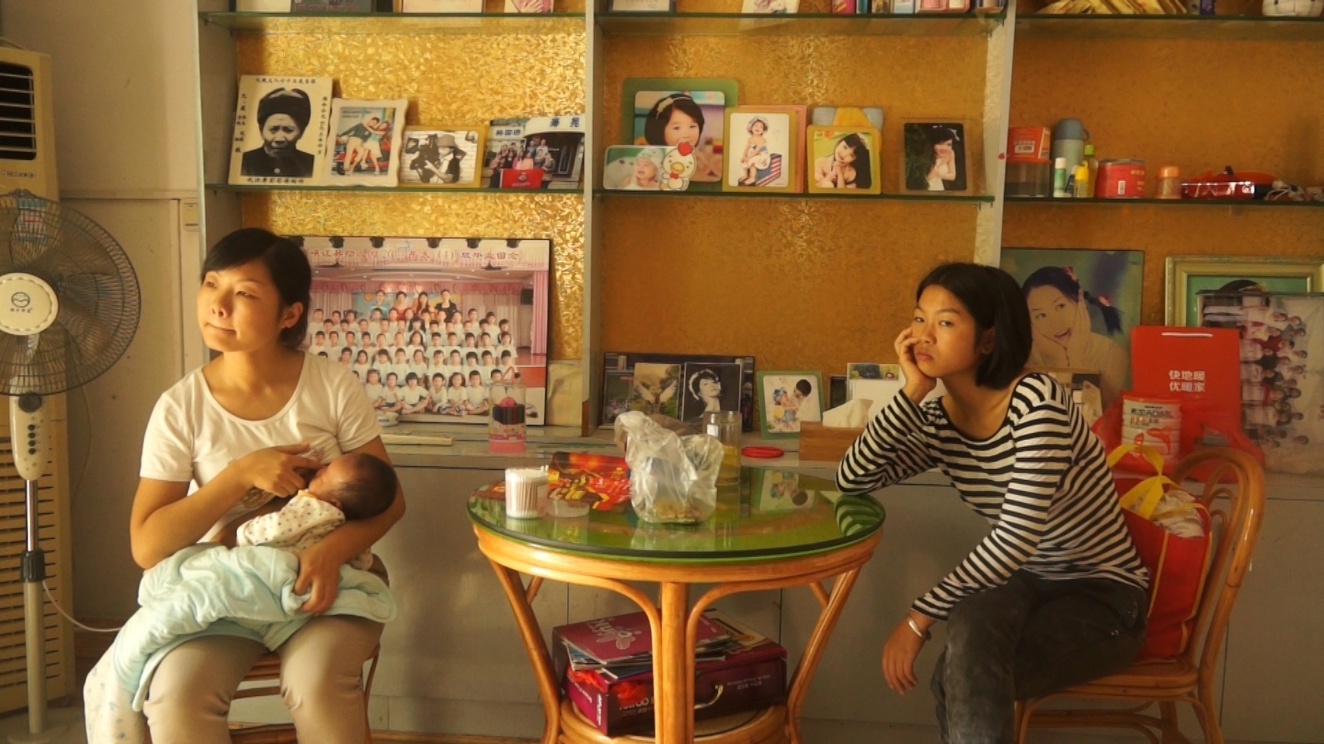 Two adults sit at a cafe. One person at the left is also carrying a baby who they are feeding. The person on the right leans their head on their hand against a table, looking bored.