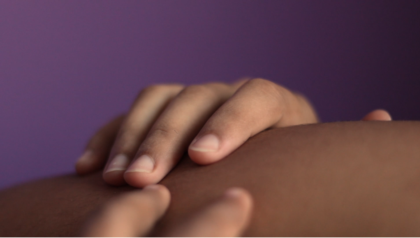 Still from "À beira do planeta mainha soprou a gente" shows a close up of a hand resting on someone's skin. The exact body part is unidentifiable but a deep purple background contrasts with the brown skin tones.