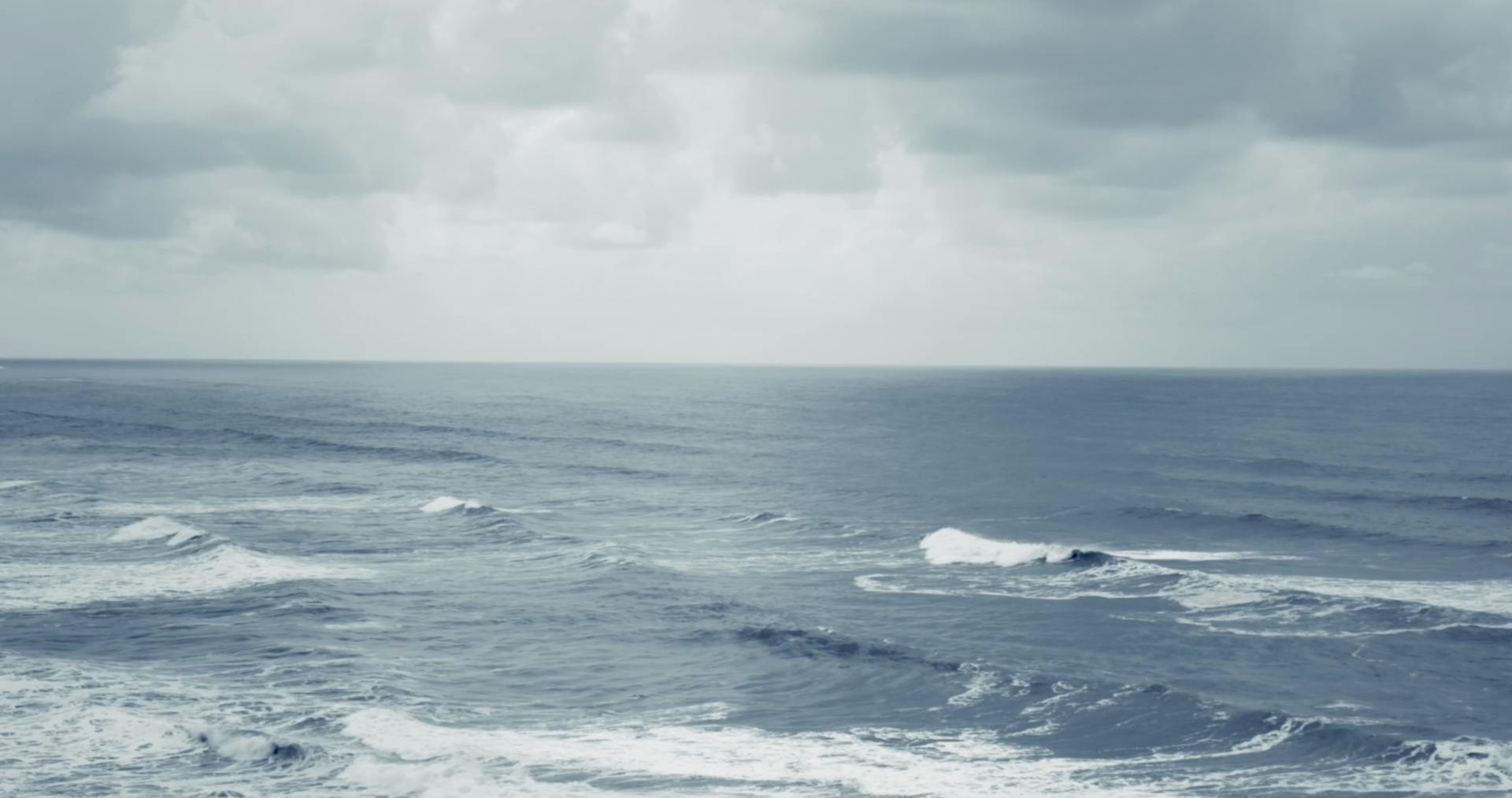 Still from the film małni shows the ocean on a cloudy day. Waves can we seen rolling towards the shore as dark clouds gather above the water.