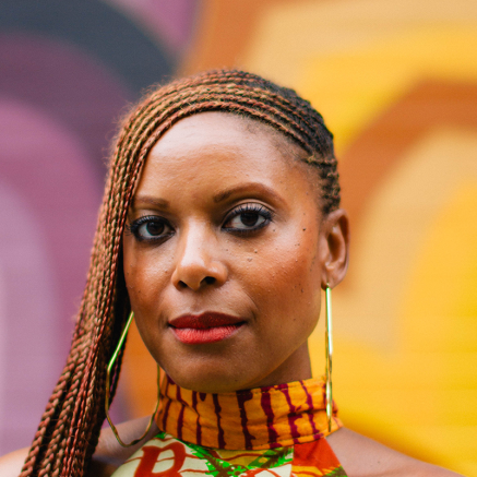 A photo of a Black person standing in front of a colorful background, their har is braided and parted to the left side. They look serenely at the camera.