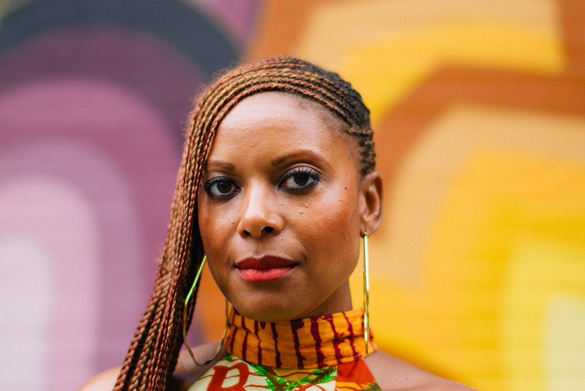 A photo of a Black person standing in front of a colorful background, their har is braided and parted to the left side. They look serenely at the camera.