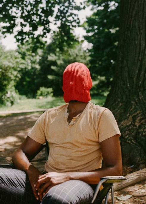 A Black person sits in a chair outside, in front of a tree, their face covered by an orange/green beanie pulled down to their kneck.