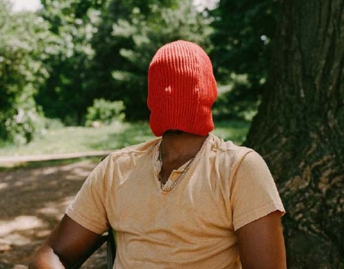 A Black person sits in a chair outside, in front of a tree, their face covered by an orange/green beanie pulled down to their kneck.