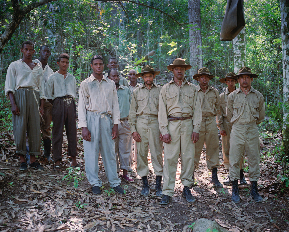 Image Description: A group of twelve men, predominantly dressed in khaki and tan shirts, pants, and wide-brimmed hats, stand in a forested area and look into the camera.