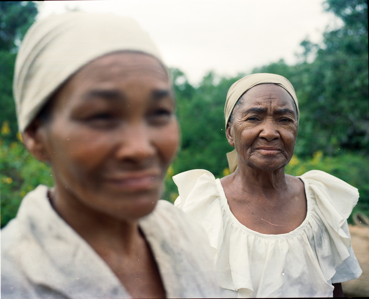 Image Description: A photo of two older women wearing light tan clothing and head coverings. The camera focuses on one woman in the foreground who looks directly at it, while the woman closest to the lens is out of focus.