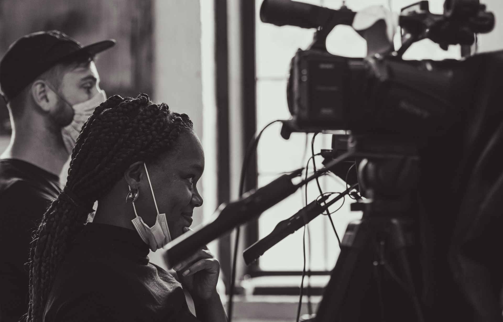 Two people stand behind a camera, looking on at a scene they are presumably recording. They both wear face masks though the person in front, who is Black, has pulled their face mask down to smile at what they are looking at to the right off-screen. The photo is in black and white.