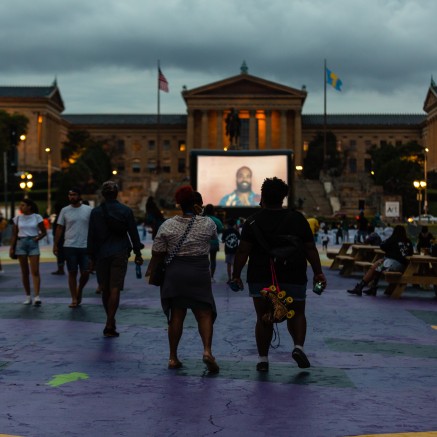 Photo of an outdoor film screening at the 10th Annual BlackStar Film Festival. A thin crowd of people walks in an open park. Small groups can also be seen sitting at picnic tables. A large columned building with two flags waving can be seen in the background, as well as a large projector screen.