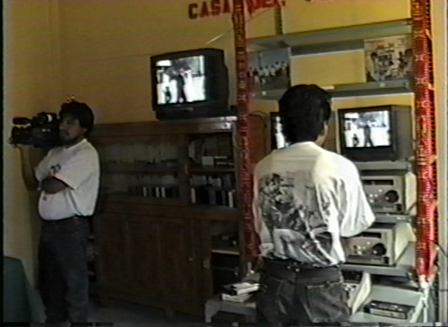 Two people inside, both wearing white t-shirts and jeans, stand around some TV screens. The one on the left holds a camera on their