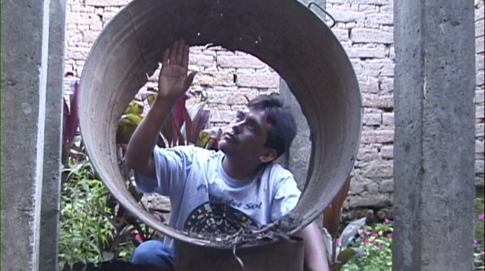 A film still shows a person with brown skin looking through a circular object, up at the interior of the object, while sitting outside with it.