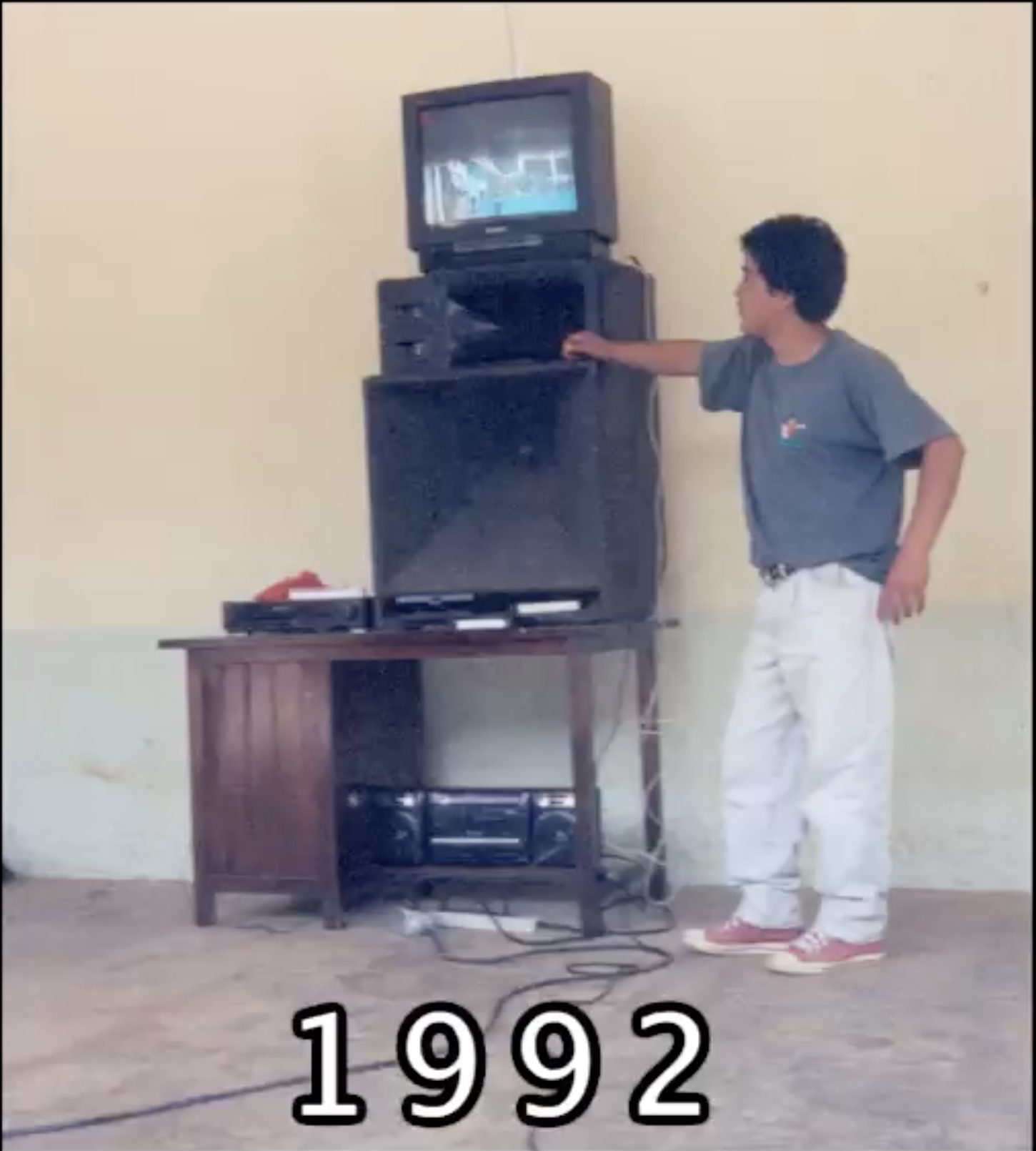 A person with brown skin stands next to a television, which sits on a media tower. At the bottom of the photo is the date, "1992".