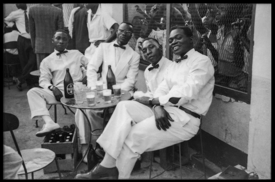 A group of Sapeurs seated in front of glasses of Primus beer, Kinshasa, ca.1955-1965.