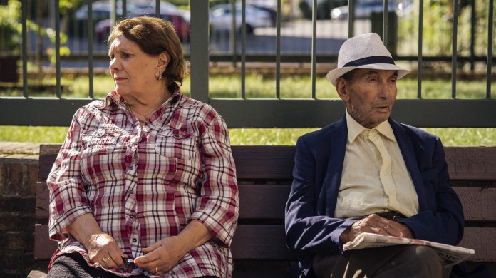 Two older people sit on a park bench facing opposite directions, but sitting close enough that they seem to know each other.