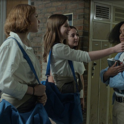 A still from the BGM music video. It shows 4 girls in school uniforms in a school hallway. Three of the girls are white, the girl opposite them is Black. One of the white girls is reaching out to touch the Black girl's girl.