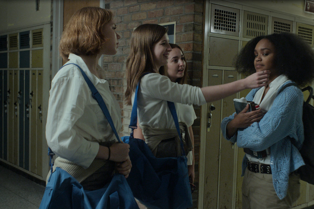 A still from the BGM music video. It shows 4 girls in school uniforms in a school hallway. Three of the girls are white, the girl opposite them is Black. One of the white girls is reaching out to touch the Black girl's girl.