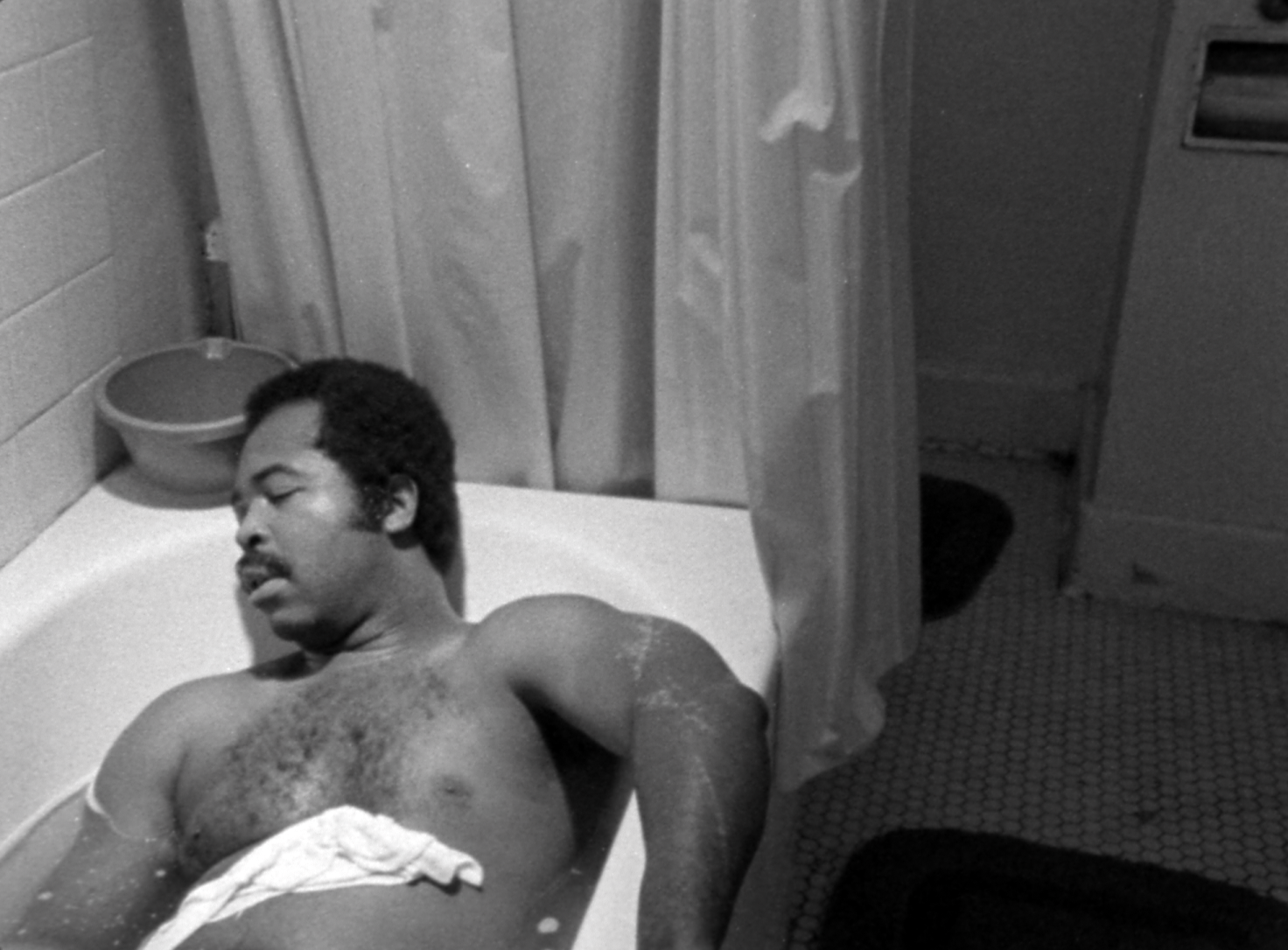 A Black person seems to be asleep in a bathtub, shirtless, shot in black and white.