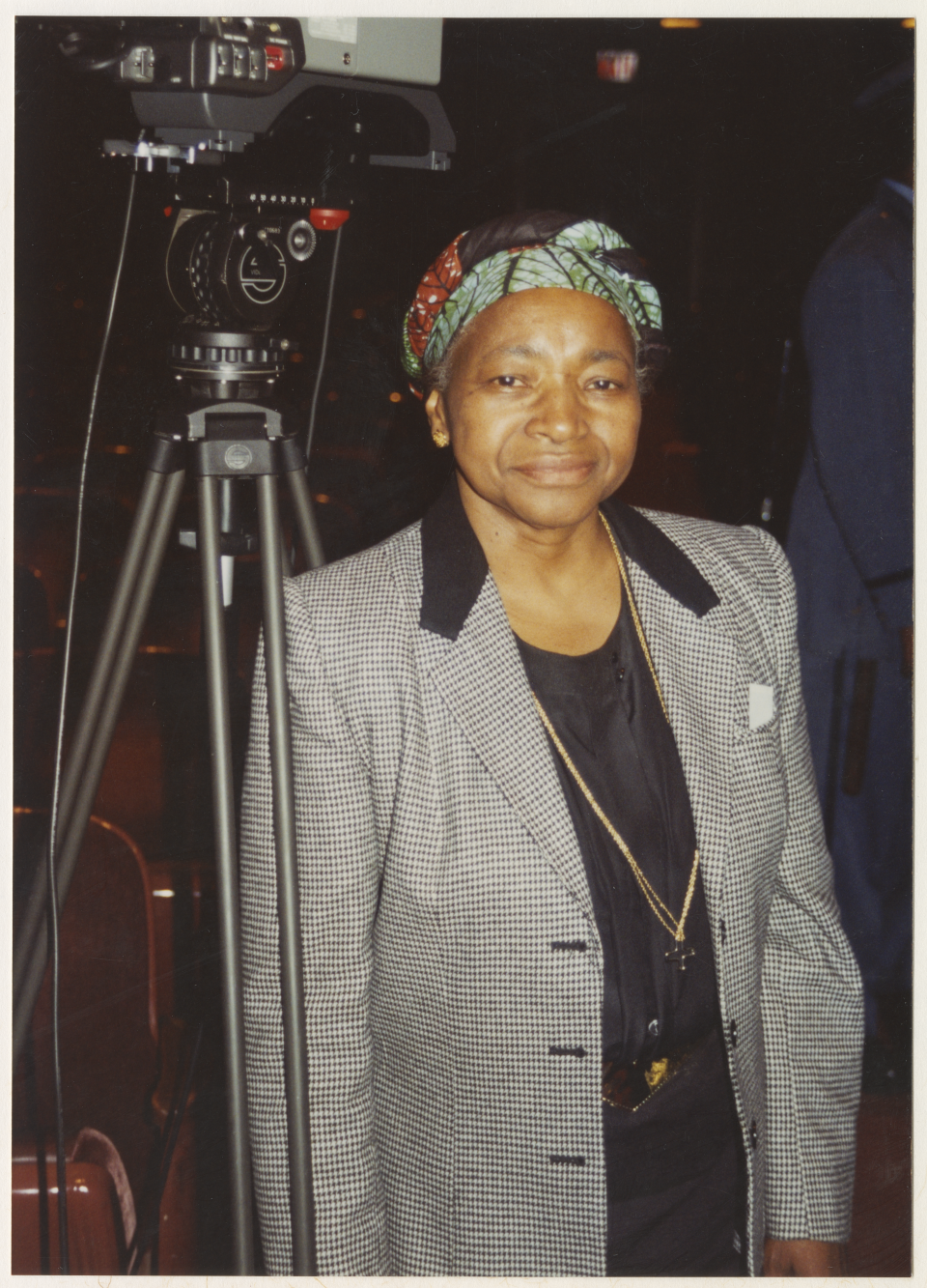 Photo of filmmaker Madeline Anderson shows her standing beside a film camera and tripod. She wears a grey blazer and a multicolored headscarf.