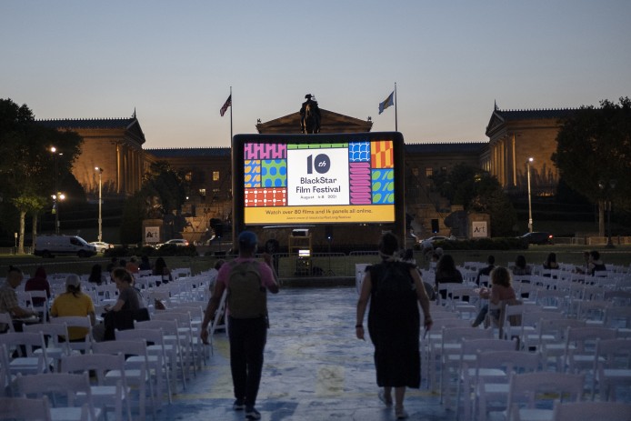 A photo of an outdoor screening during the 10th BlackStar Film Festival taken during the early evening. The screen is visible in the distance as two people walk towards it in shadow.