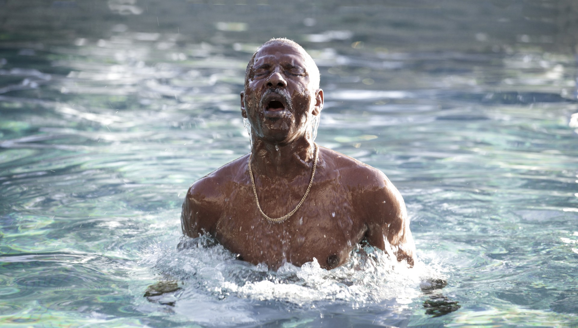 A Black person emerges from the water shirtless, wearing a gold chain, their eyes closed.