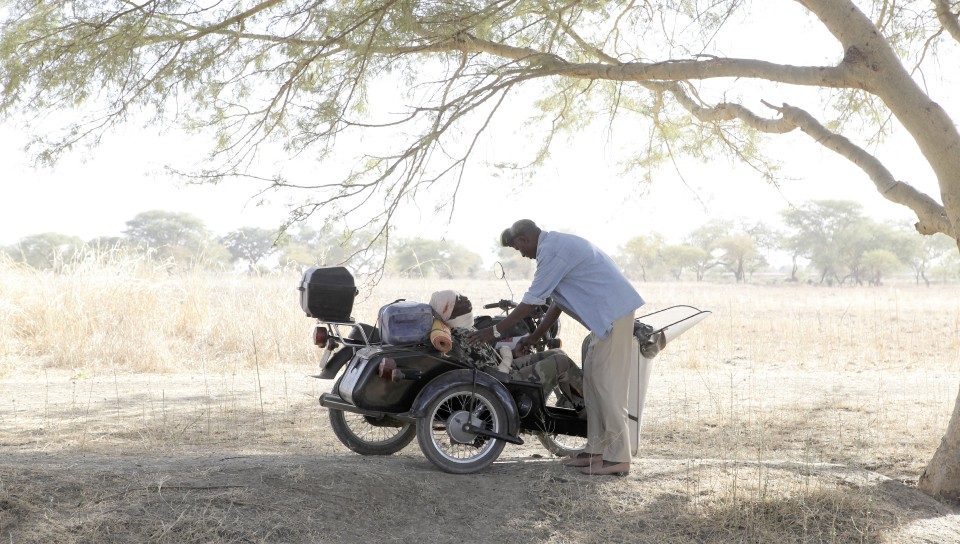 A Black person fixes a motorcycle outside under a large tree