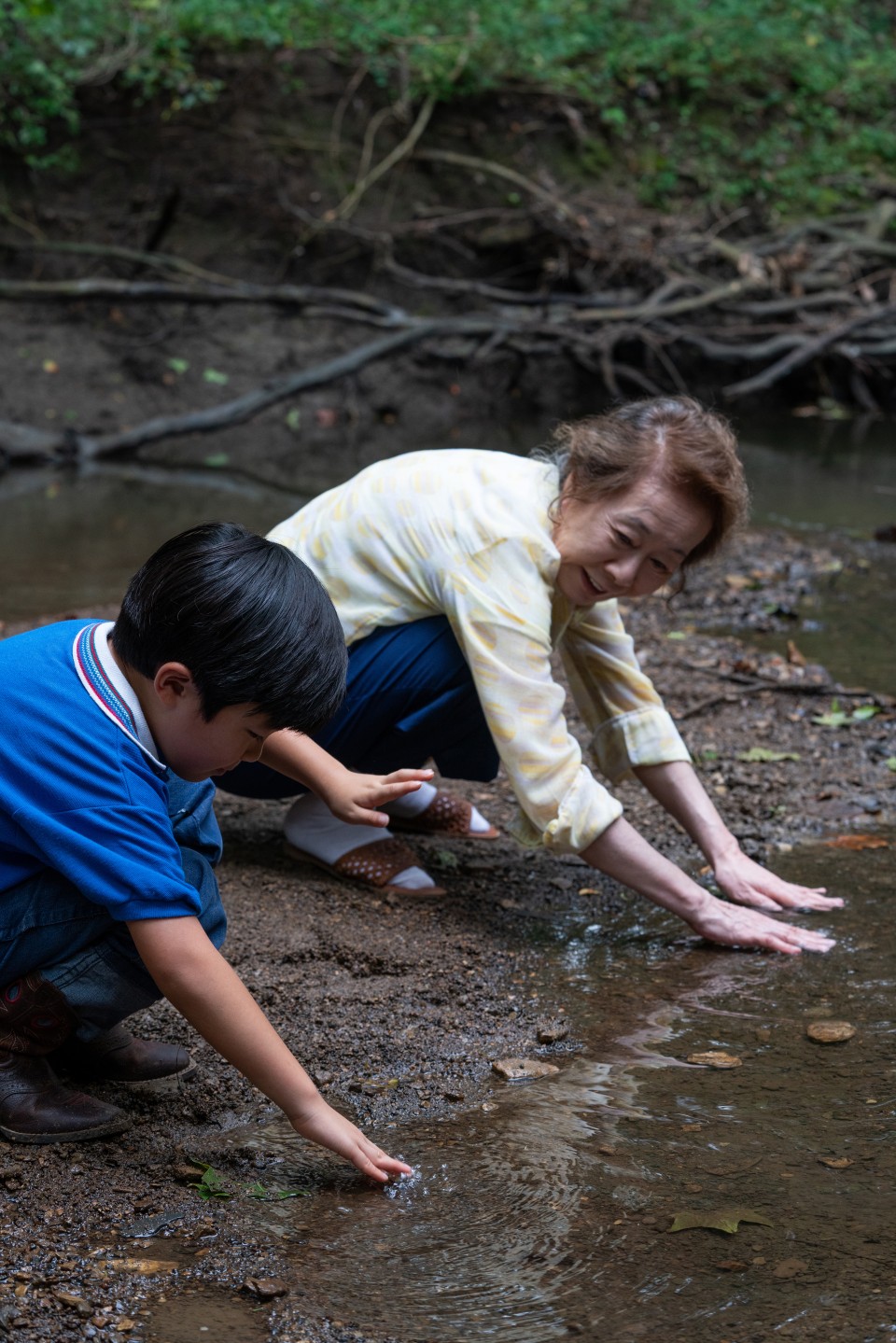 Still from the film "Minari" shows an older Korean woman and a young boy crouching at the bank of a shallow river. They place their hands in the muddy water as the older woman smiles at the child.