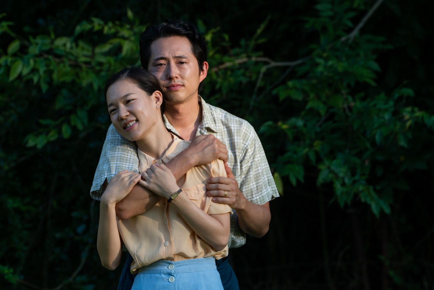Still from the film "Minari" shows a young Korean couple embracing. A man stands, wrapping his arms around a woman who leans into his body and returns his embrace. The woman smiles and looks off-screen while the man stares forward with a blank, almost worried, expression