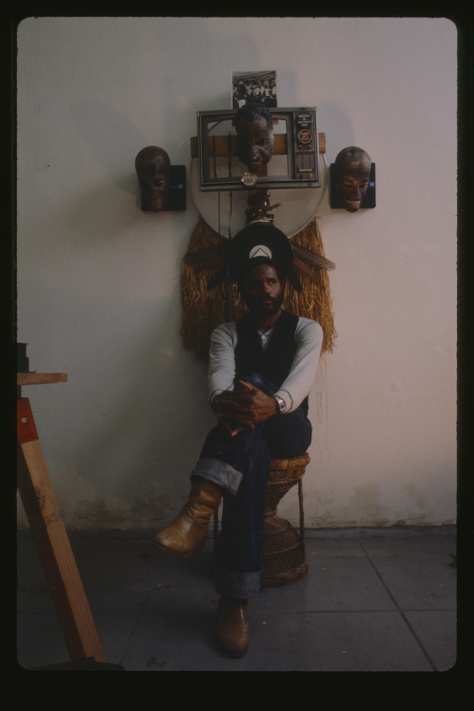 Ulysses Jenkins in studio circa 1983. The artist sits with their legs crossed in front of a complex piece of work.