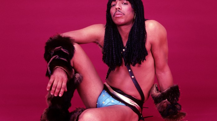 A photo of Rick James shows him posing while sitting on the floor in only underwear, his right arm drapped over his right knee, which is raised off the ground. The background is all red.