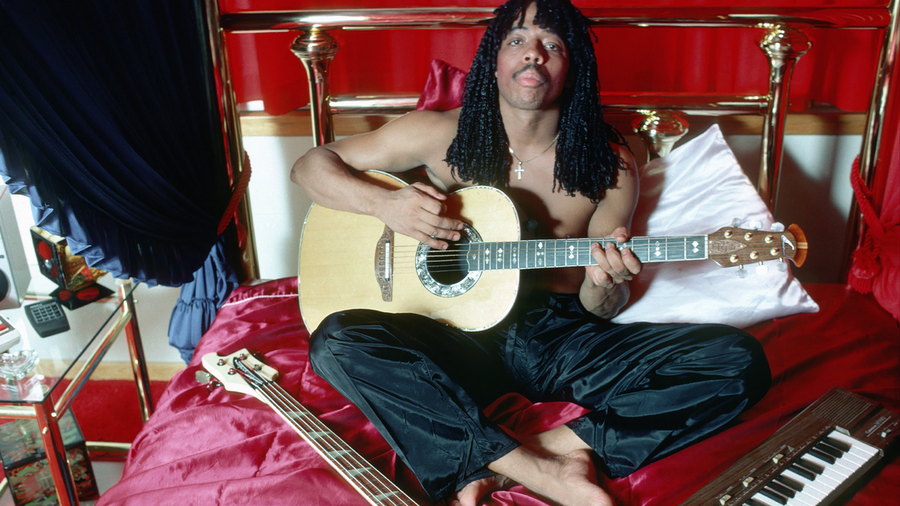 Rick James playing guitar while sitting on a bed, shirtless.