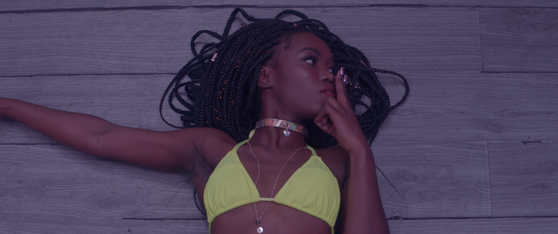 A still from She Paradise by Maya Cozier shows a Black person laying down and raising a finger to their lips while looking left, wearing a yellow-neon bikini top.