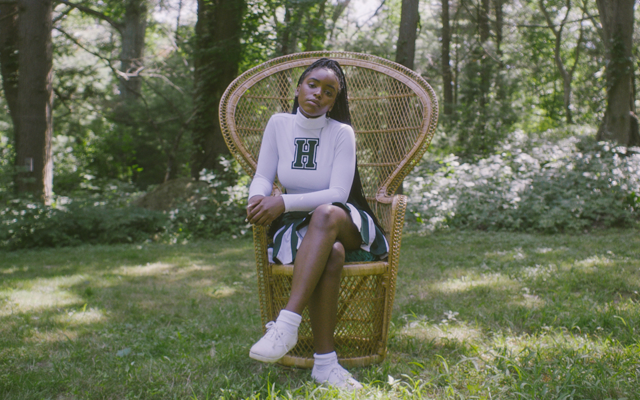A still from Selah and the Spades. A young woman wearing a green and white cheerleading uniform is sitting on a woven peacock chair. There is a forest in the background. She is the middle of the frame looking directly at the camera.