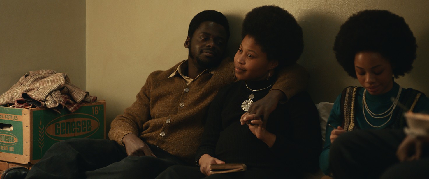 Image Descriptions: Still from "Judas and the Black Messiah" picturing three young Black people, one man and two women, sitting with their backs against a wall. Both women have large afros and wear silver necklaces. The man's arm is wrapped affectionately around the shoulders of one of the women. They appear to be a couple.