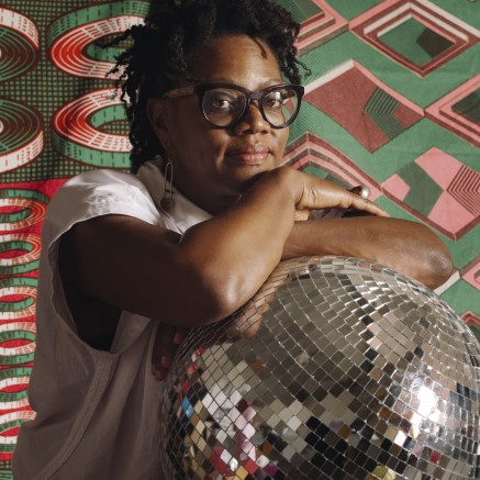 A photo of Cauleen Smith, who is a Black person wearing black-framed glasses, arms-crossed over a disco ball, in front of a colorful print.