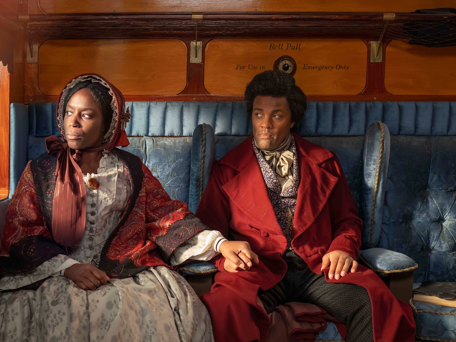 Two Black people sit inside a carriage, dressed in period attire. They both look out the window on the left pensively, the person on the far left - who wears a bonnet and floral dress - has their left hand on the right hand of the person on the right, who is wearing a red overcoat and appears to be portraying Frederick Douglass.
