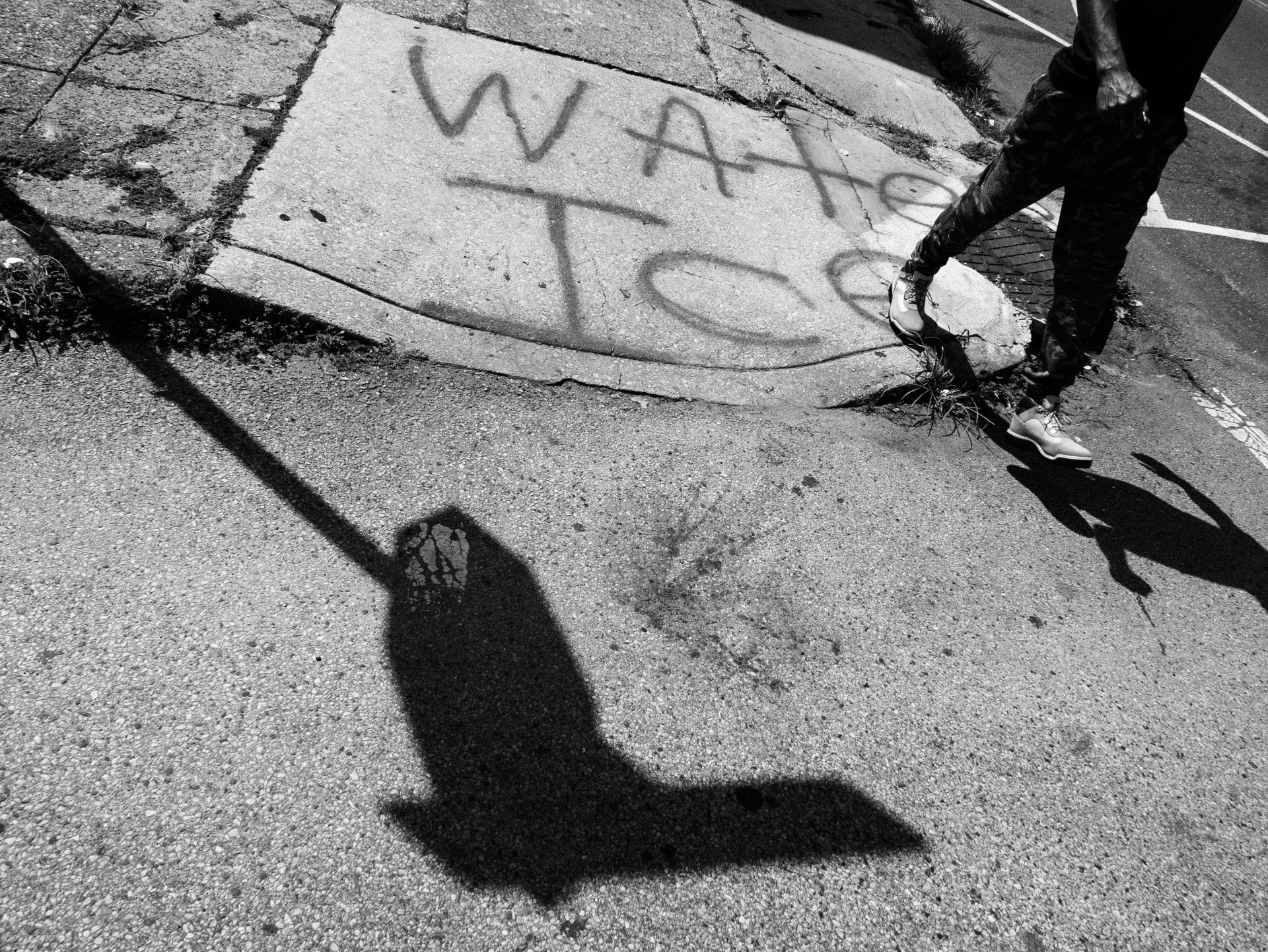 A black-and-white photo of a street corner. The photo is shot from above, it shows a sidewalk curb, a stop sign shadow, and a man walking. Graffitied on the sidewalk are the words "water ice".