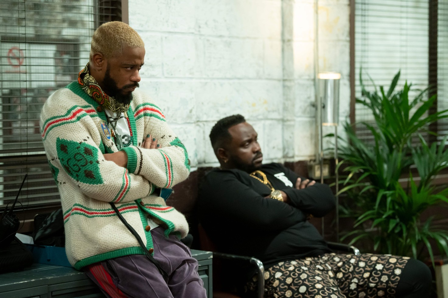 Two Black men (LaKeith Stanfield and Bryan Tyree Henry) stare skeptically offscreen. LaKeith leans against a wall in the foreground with his arms crossed against his chest, while Bryan sits in a chair, arms also crossed.