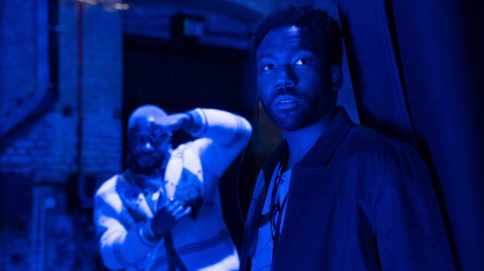 Two Black men stand facing the viewer in a dark, blue-lit space. Donald Glover stands in the foreground, looking off frame, while LaKeith Stanfield stands in the background, with a hand framing his left eye.