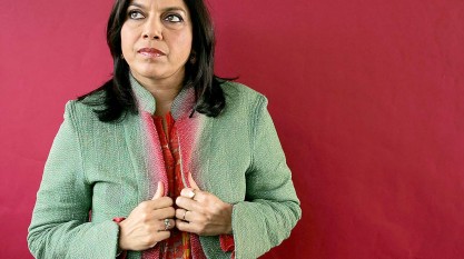 A headshot of Mira Nair, she is standing in front of a red wall. She is wearing a green and red coat and she is looking off to the side with a contemplative expression.