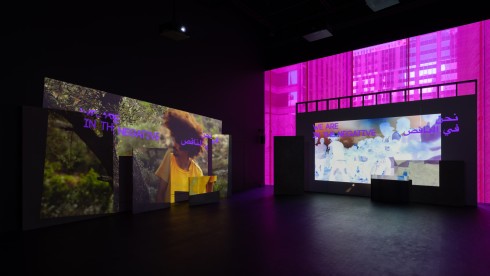 Installation view of Basel Abbas and Ruanne Abou-Rahme: May amnesia never kiss us on the mouth. There are two large projections. The one on the left has an image of a person with big curly hair and a yellow t-shirt, they are in a forest-like setting. The text on top of the image says "WE ARE IN THE NEGATIVE" in English, beside that is Arabic text that presumably says the same. The projection on the right is a blown-out image of a group of people, their silhouettes are a bright white. The text on the image says "WE ARE IN THE NEGATIVE" in English, beside that is Arabic text that presumably says the same.