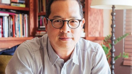A headshot of Jeff Chang. He is wearing black-rimmed glasses and a pin-stripped button down shirt. He is looking at the camera with a slight smile on his face. Behind him is a bookcase.