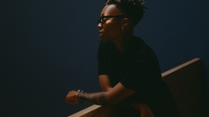 A portrait of Kya Lou, a Black person, who looks down over a bannister which they are leaning on (wearing a black short-sleeved shirt). They also wear black framed glasses and behind them is a deep blue-colored wall.