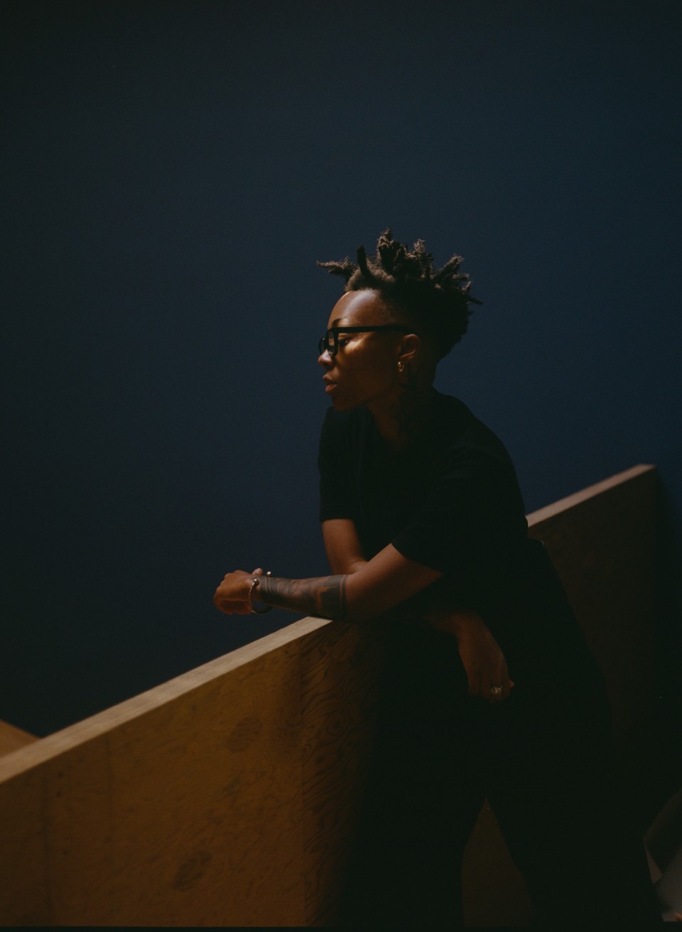 A portrait of Kya Lou, a Black person, who looks down over a bannister which they are leaning on (wearing a black short-sleeved shirt). They also wear black framed glasses and behind them is a deep blue-colored wall.