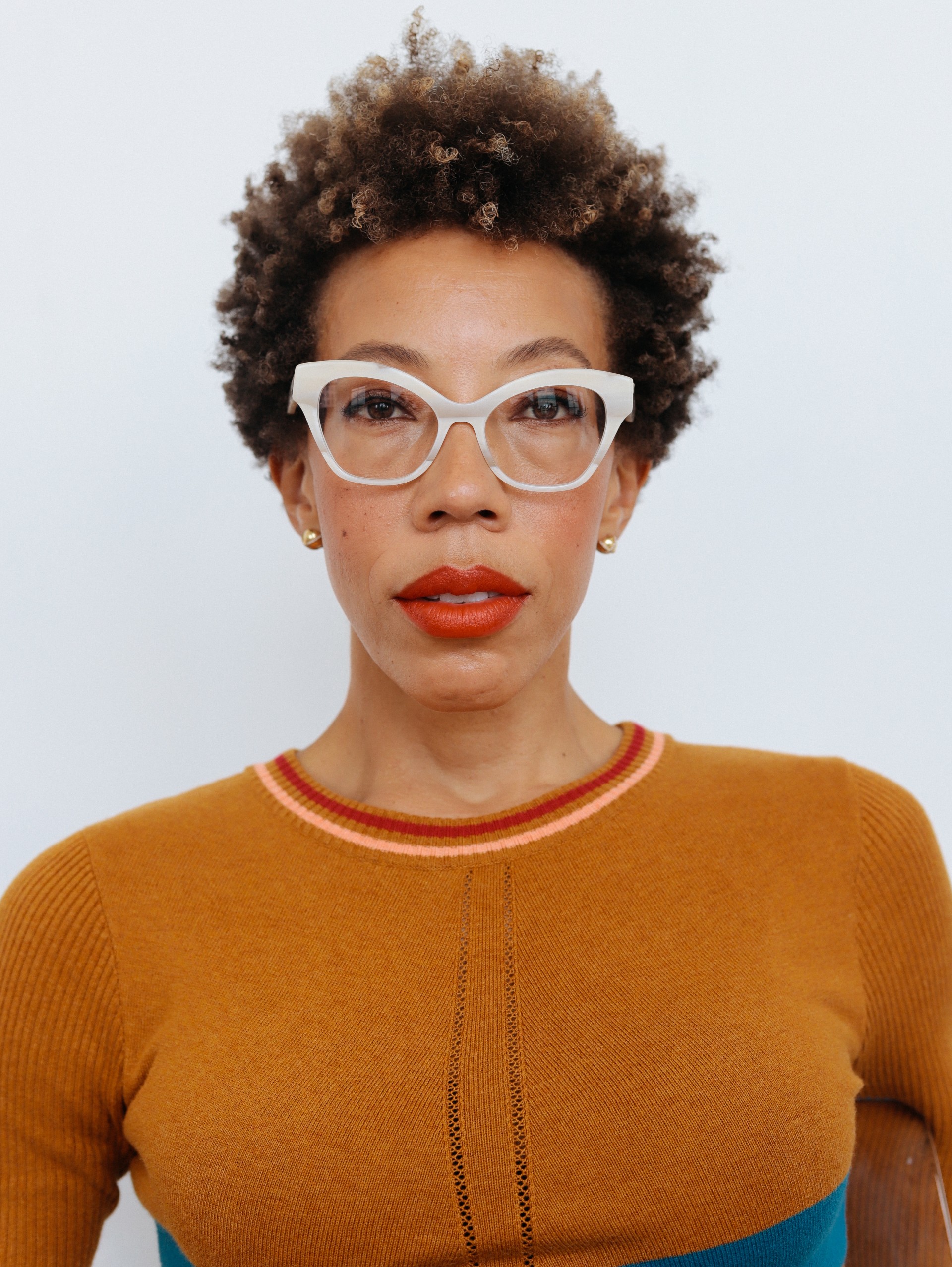 A headshot of Amy Sherald. She is wearing white-framed glasses and a tan sweater. She is looking directly at the camera with a thoughtful expression.