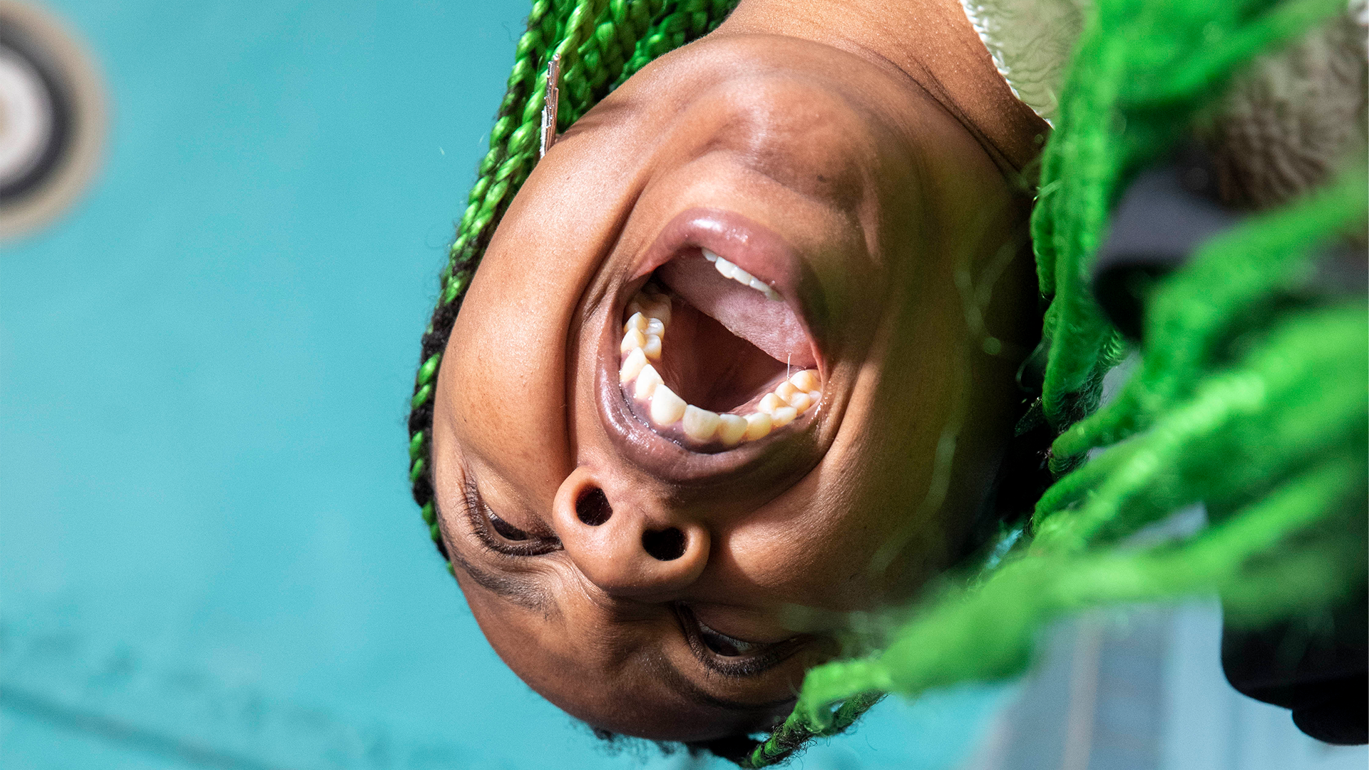 From beneath we look up at the image of Martine Syms pressing an apartment call button and apparently yelling. They have bright green braids and above them is a blue ceiling.