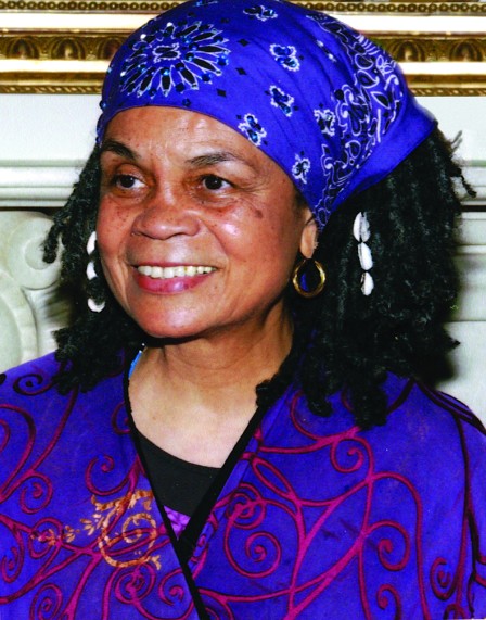A headshot of Sonia Sanchez. She is wearing a purple suit jacket. Her hair is loc'd and pushed back with a purple bandana. She has cowrie shells in her hair and she is smiling.