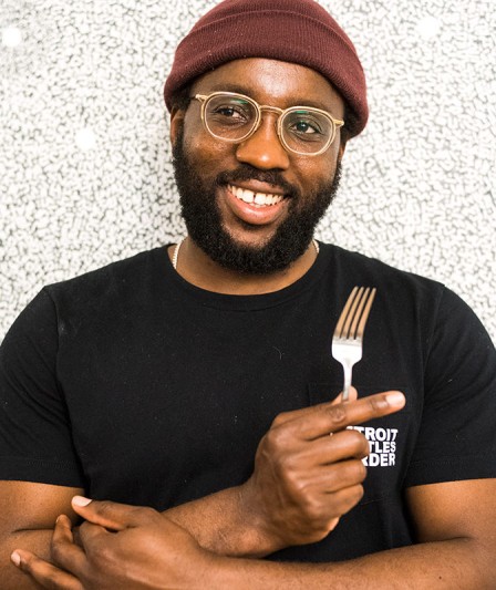 A headshot of Tunde Wey, he is wearing a burgundy beanie, a black shirt and has on clear glasses. He is holding up a fork, smiling, and looking slightly towards the left.