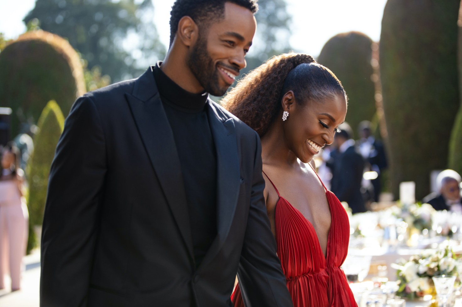 Two Black people are smiling and happy, one wears a black suit with short hair and a beard, the other - who appears to be holding their hand - wears a red dress and their long hair is tied in a ponytail. They are the characters Lawrence and Isse from Insecure.