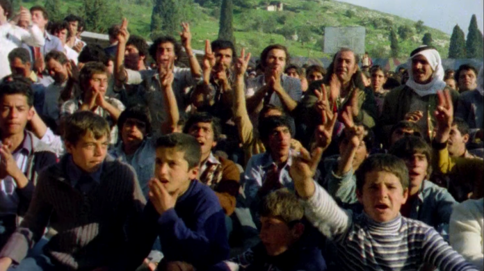 Still from Yom al-Ard (2019), dir. Monica Mauer, showing a crowd of children in Palestine raising their hands and cheering. Behind them are green hills.