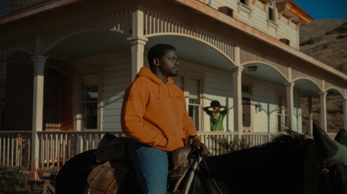 A still from Nope shows Daniel Kaluya's character sitting on a horse, looking off to the right, wearing a bright orange sweatshirt. In the background, on the porch of an old house, is Keke Palmer's character looking exasperated.